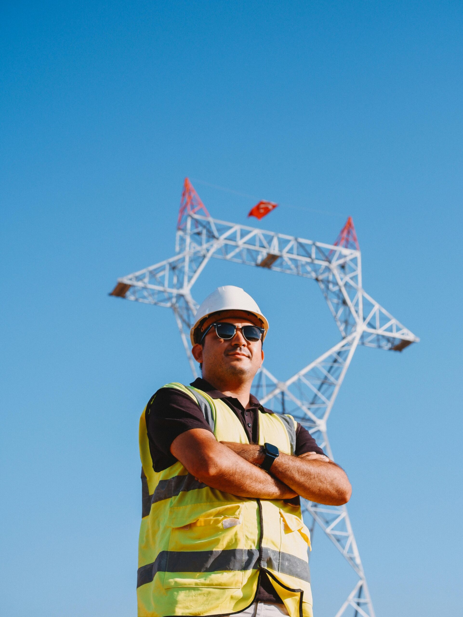 Confident engineer in safety gear stands by a power transmission tower against a clear blue sky.